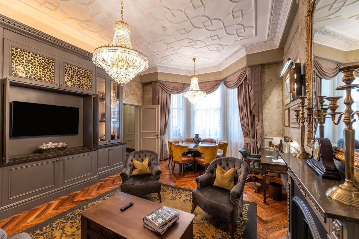 A luxurious living room with a large flat-screen TV, two armchairs with cushions, a wooden coffee table, a chandelier, and a dining area with a table and chairs.
