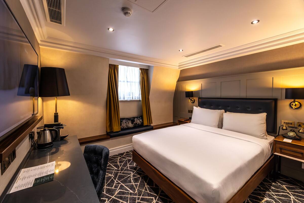 A hotel room with a large bed, a desk with a lamp, a chair, a window with curtains, and a wall-mounted TV.