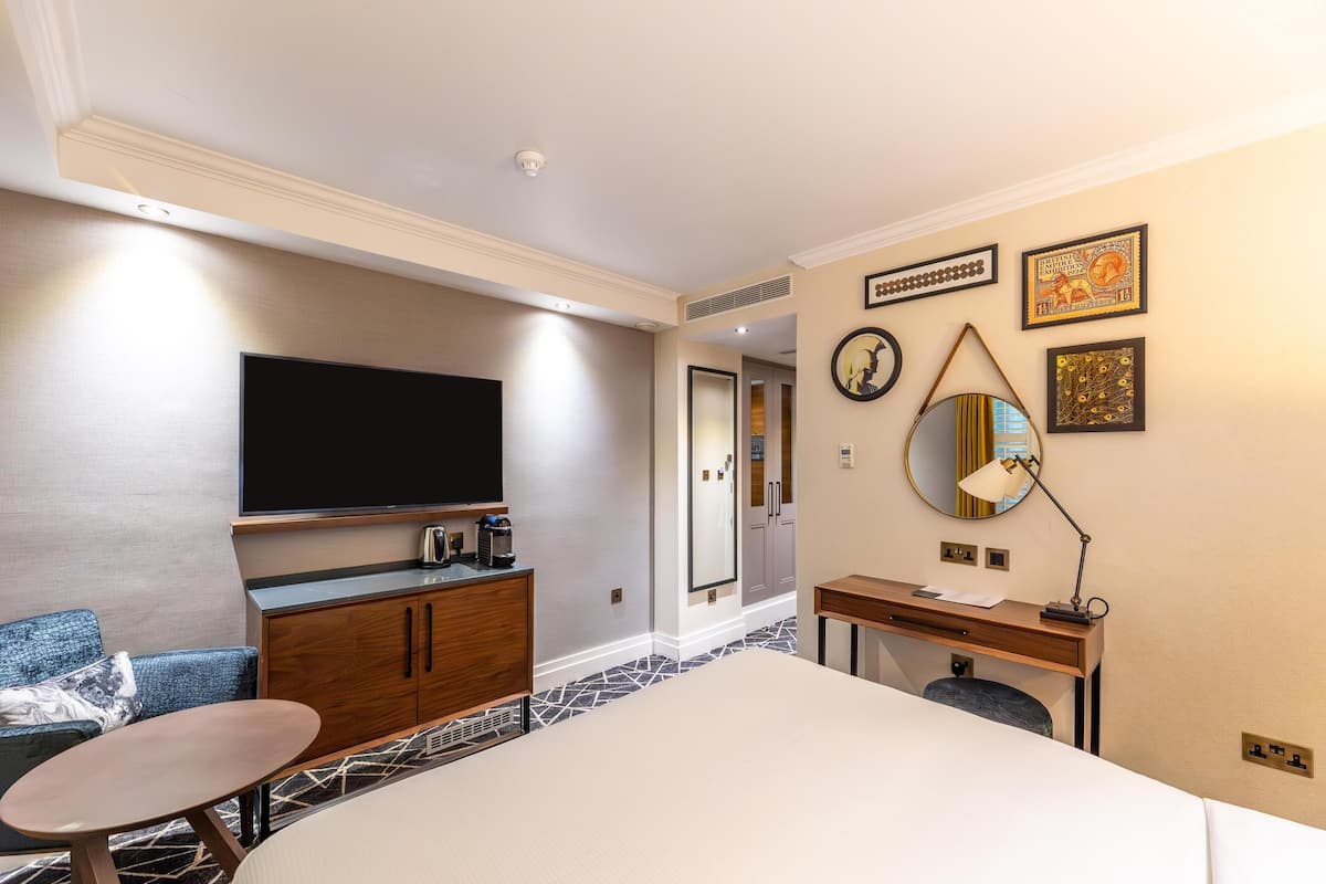 A hotel room with a bed, a TV, a desk, a chair, a mirror, and artwork on the wall.