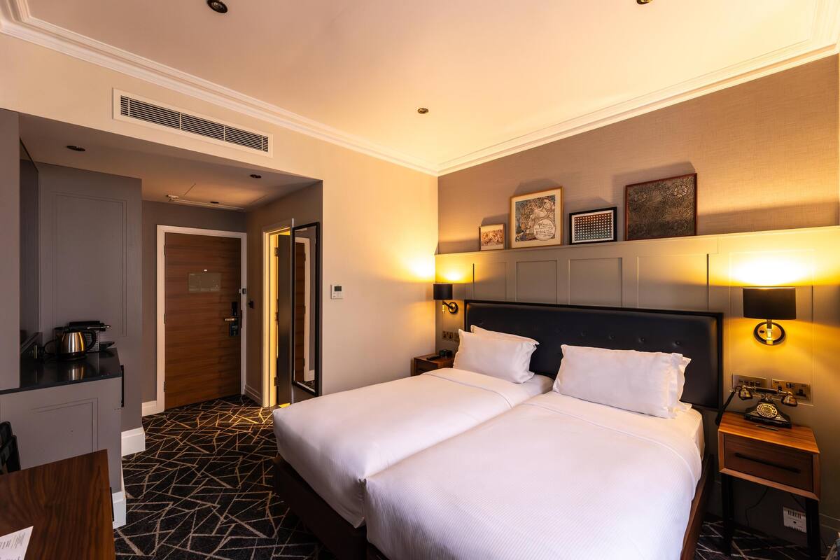 A hotel room with a double bed, bedside tables, a desk with a kettle, and a patterned floor.