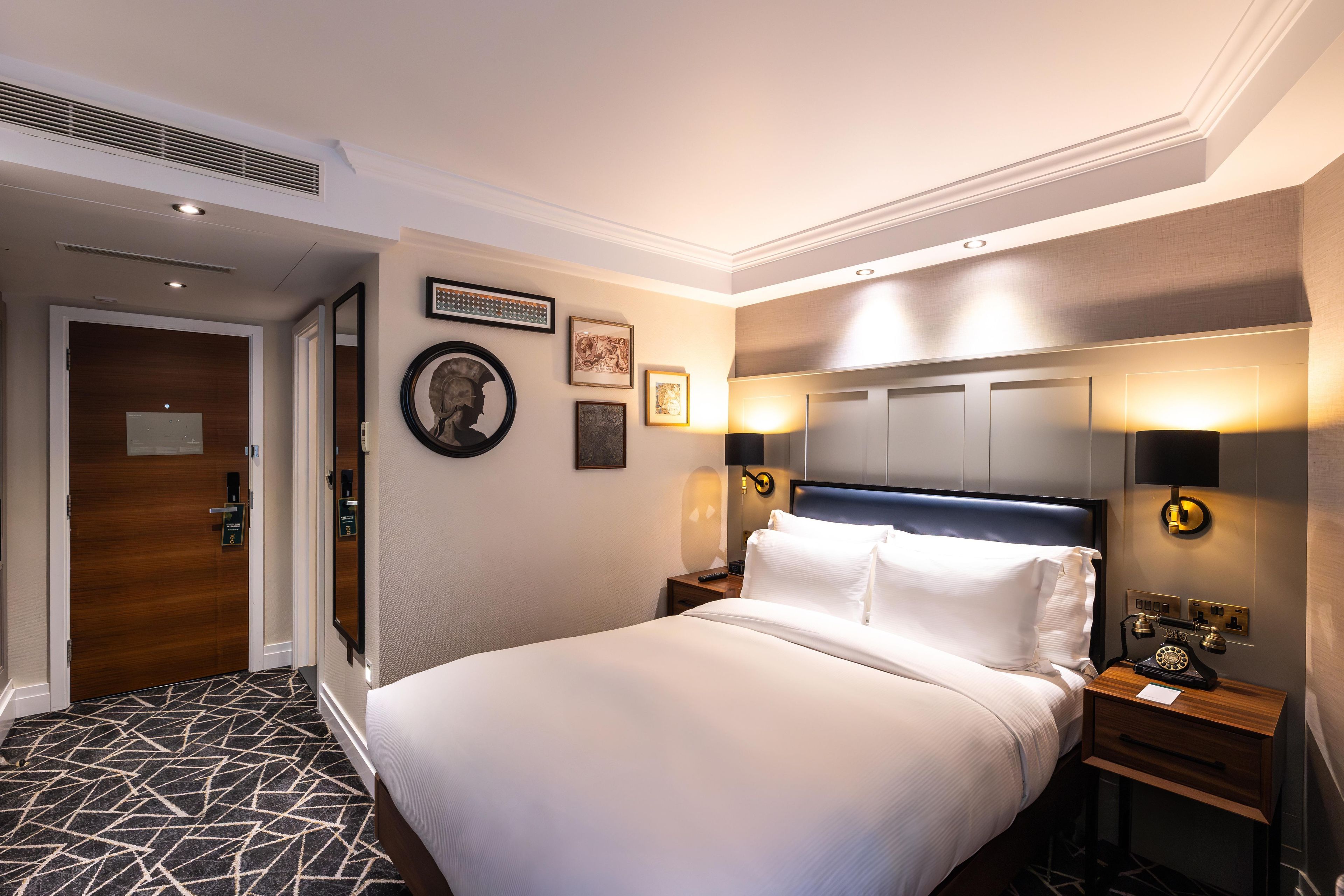 Une chambre d’hôtel avec un grand lit, une table de chevet avec une lampe, un téléphone fixé au mur et des œuvres d’art encadrées.