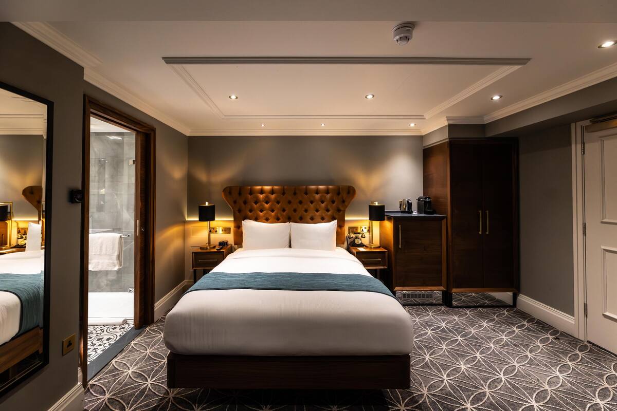 A hotel room with a large bed, a wooden headboard, a nightstand with a lamp, a wardrobe, and a bathroom visible through an open door.