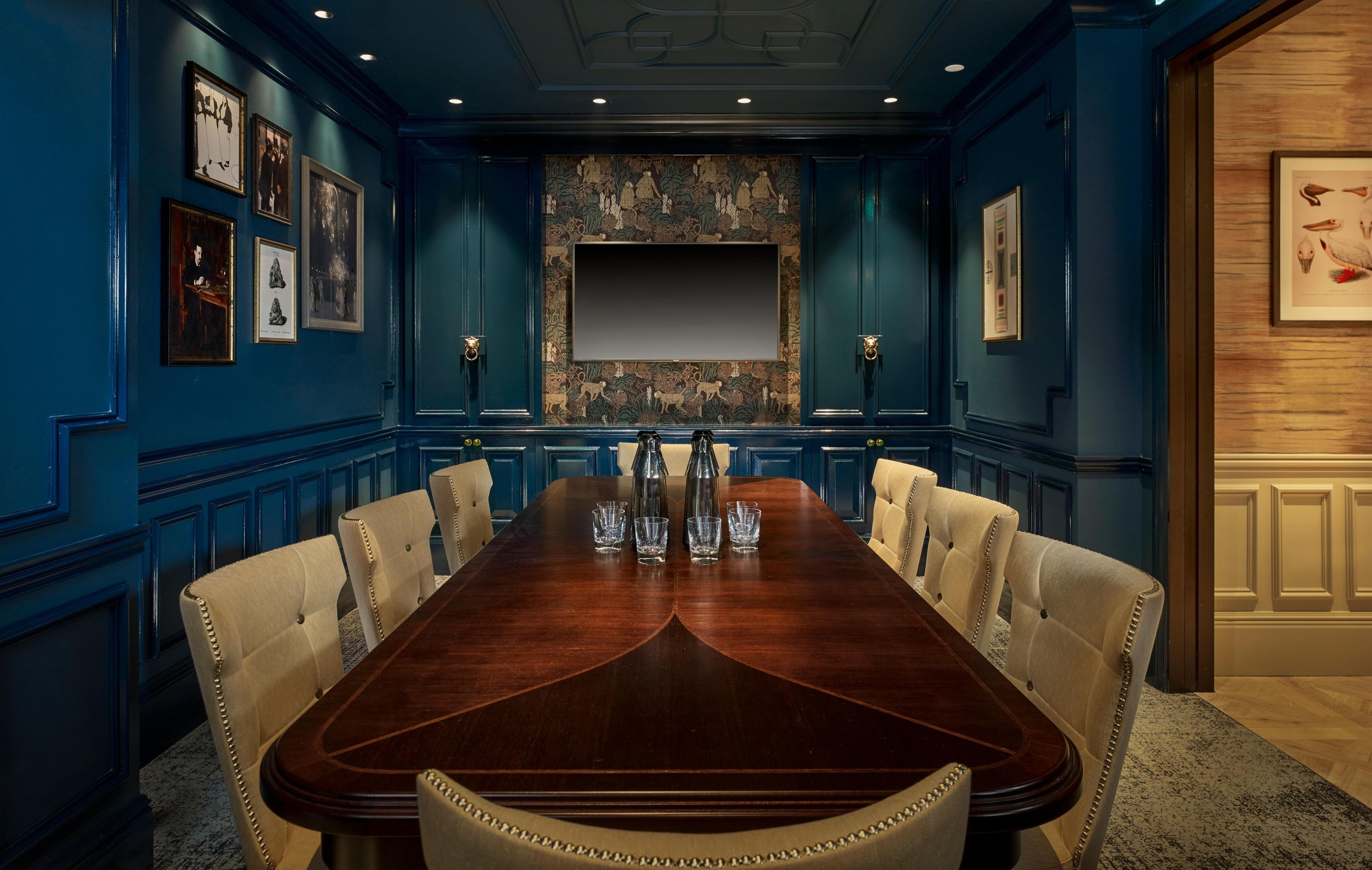 A conference room with a long wooden table, beige upholstered chairs, framed pictures on the walls, and a large blank screen at the front.