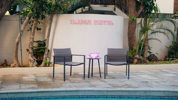 A pool area with two chairs and a small table, a sign reading "Ilima Hotel", and surrounding greenery.