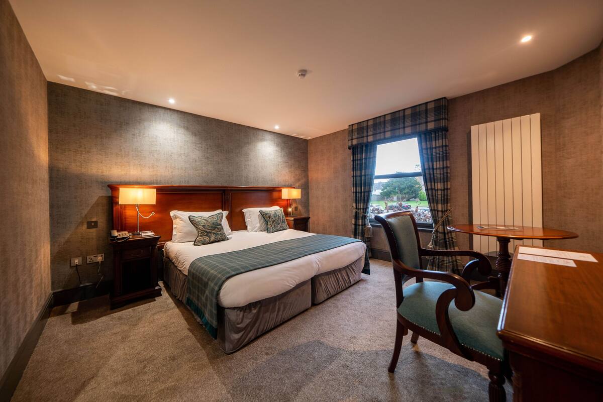 Ein Hotelzimmer mit einem großen Bett, Nachttischen, einem Schreibtisch und einem Fenster mit Vorhängen.