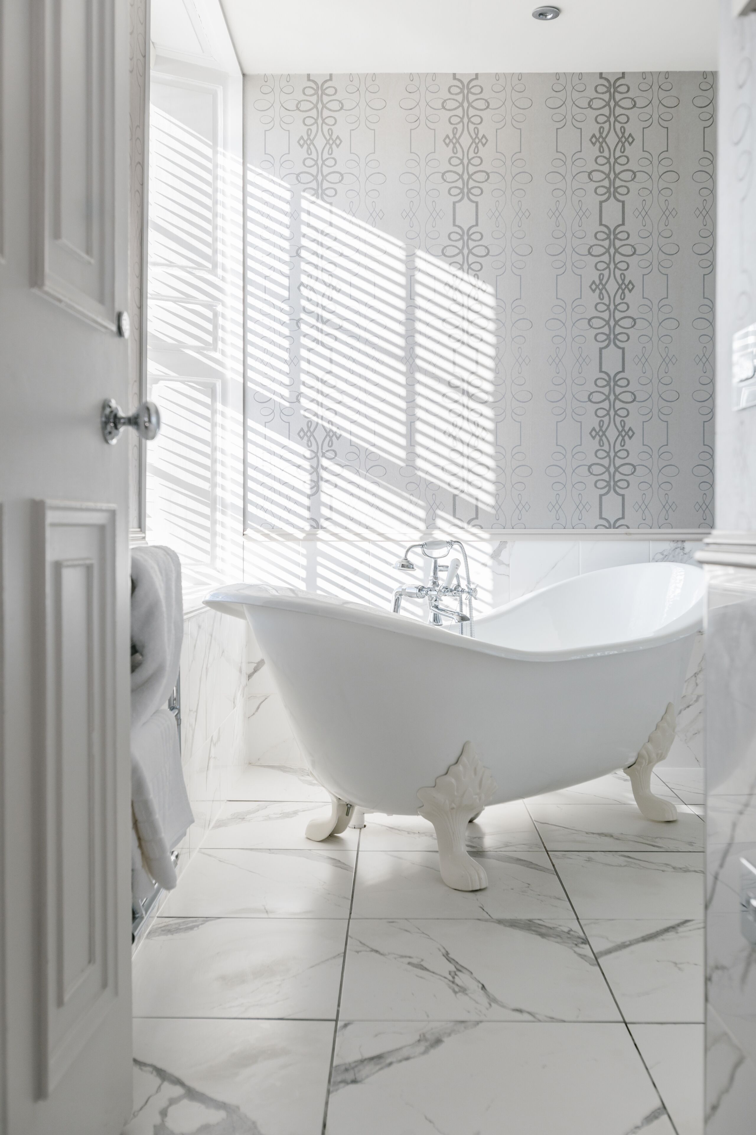 Un baño con una bañera independiente, azulejos de mármol blanco y papel tapiz estampado.