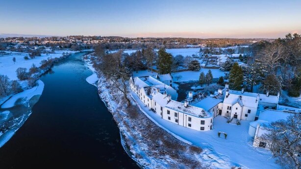 Une propriété riveraine enneigée, avec un long bâtiment attenant à la rivière, dotée de nombreuses fenêtres et d’une façade symétrique.