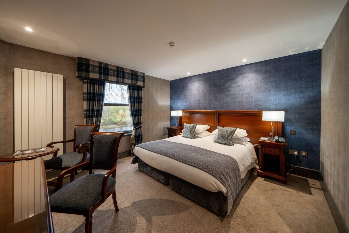Ein Hotelzimmer mit einem großen Bett, zwei Nachttischen, einem Schreibtisch mit Stuhl und Blick ins Freie durch ein Fenster.