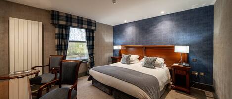 A hotel room with a large bed, two bedside tables, a desk with a chair, and a view of the outdoors through a window.