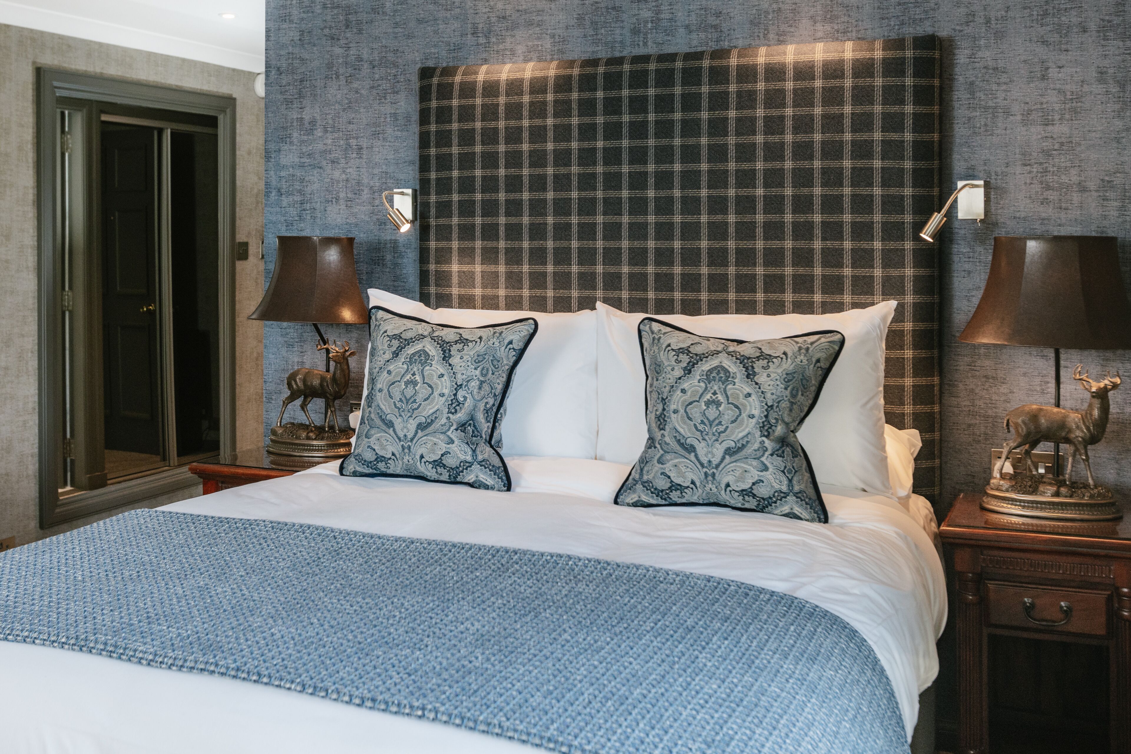A neatly made bed with a blue blanket, two matching pillows, and a headboard with a plaid pattern. There are two nightstands with lamps and a deer statue on each.