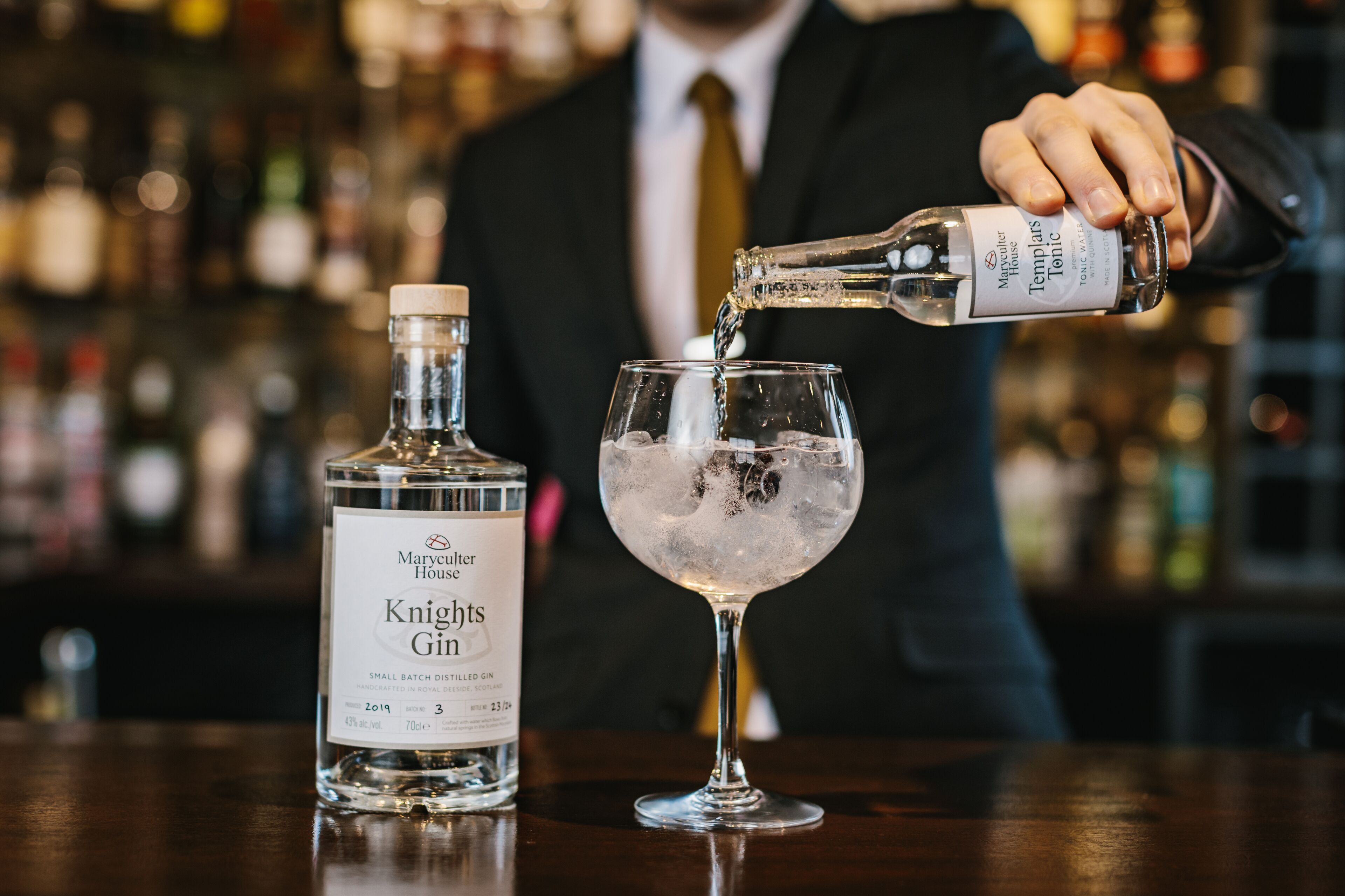 A bartender pouring a drink from a bottle labeled "Marcy Cutter House Knight's Gin" into a glass with ice.