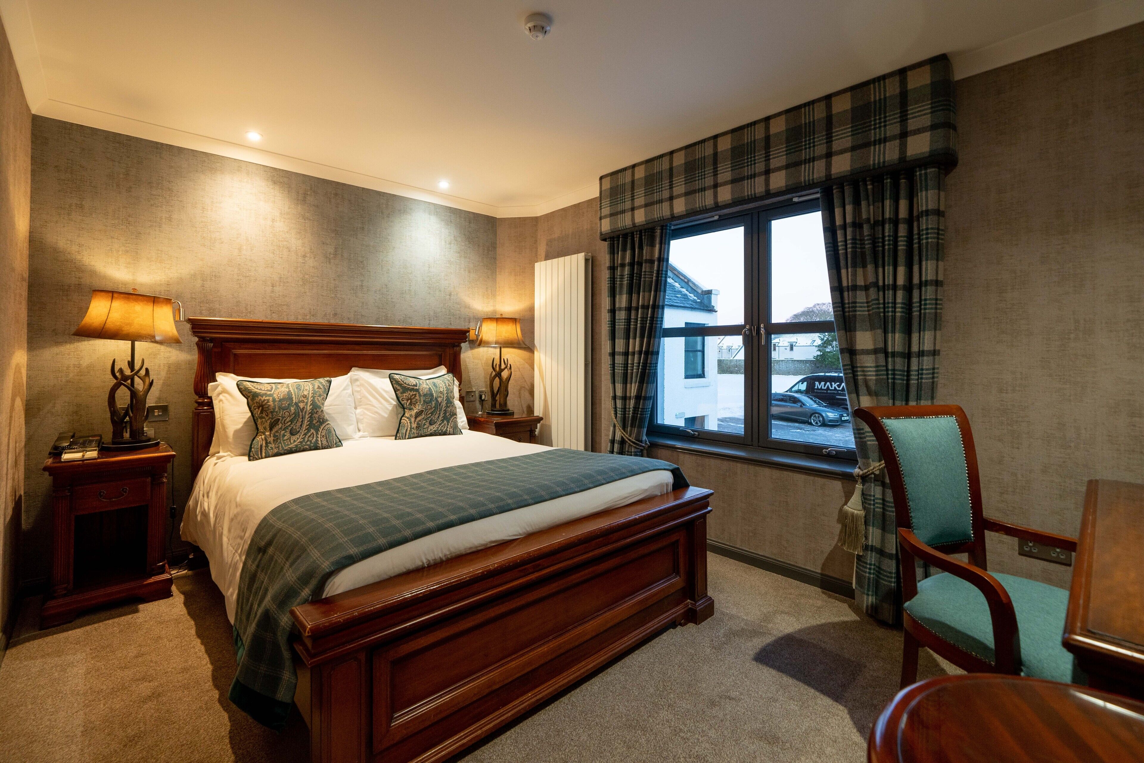 Une chambre d’hôtel avec un grand lit, deux tables de chevet avec des lampes, une chaise, une fenêtre donnant sur une voiture et un rideau à carreaux.
