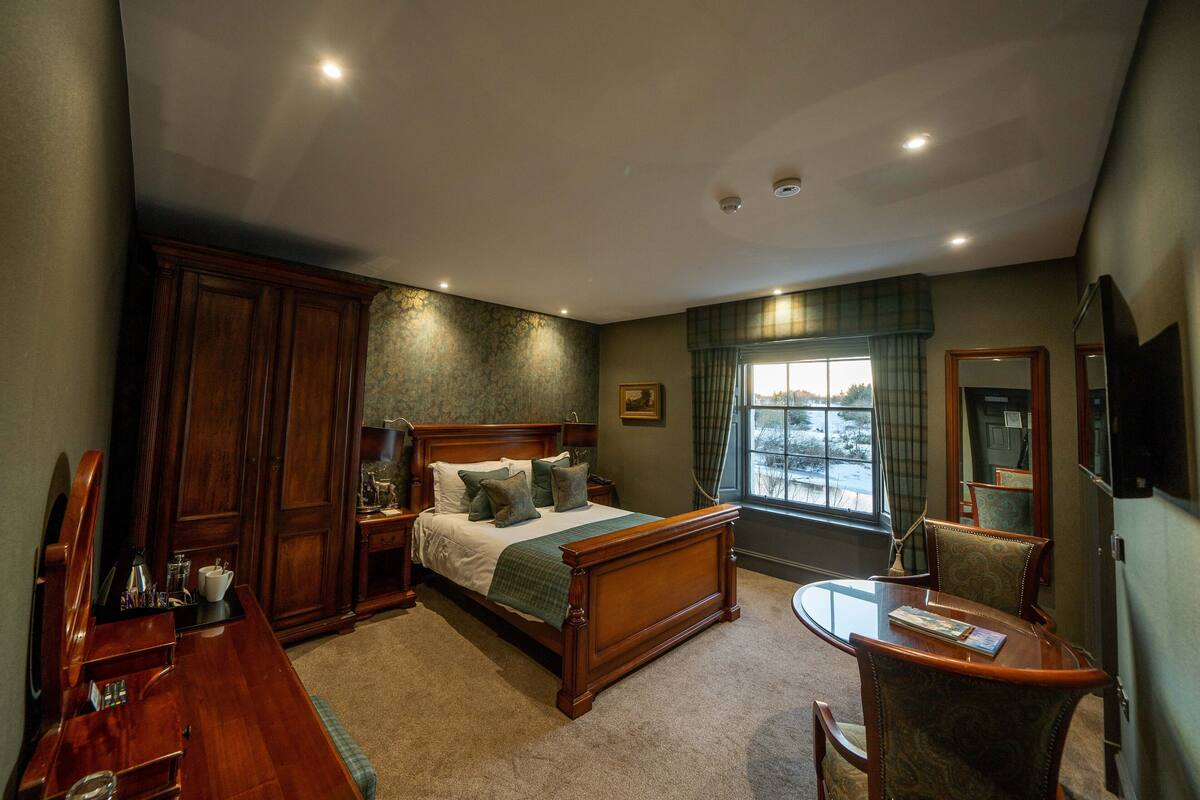 Ein Schlafzimmer mit einem hölzernen Bett, einem Schreibtisch und einem Stuhl. Ein Fenster bietet Blick ins Freie.