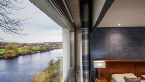 Une chambre d’hôtel avec une grande fenêtre donnant sur une rivière, un lit avec des oreillers, une table de chevet avec une lampe et un mur sombre à la texture marquée.
