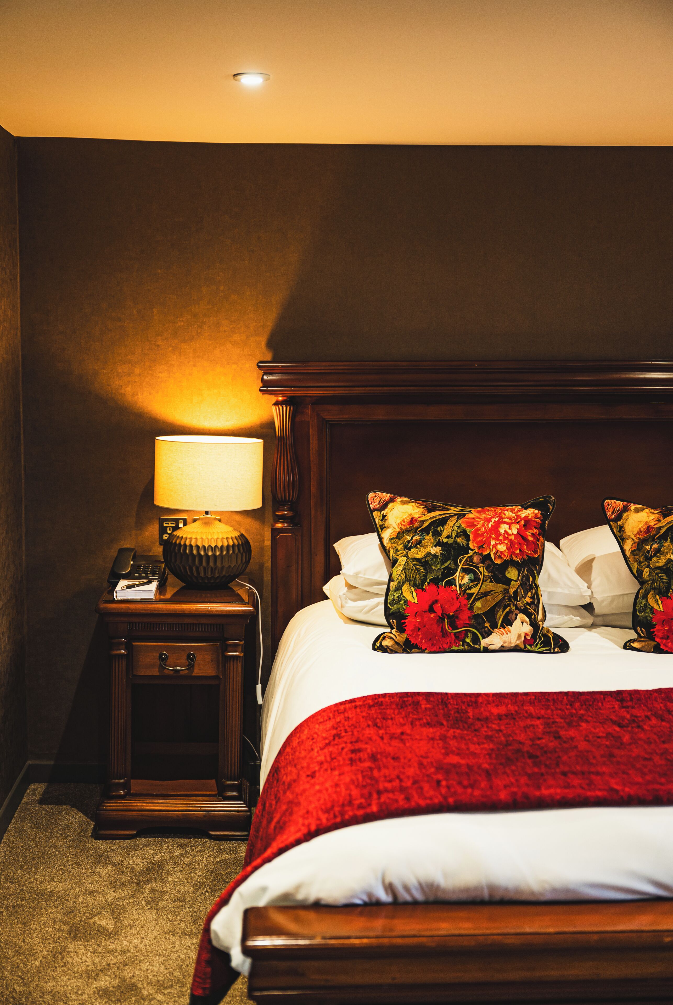 A hotel room with a wooden headboard, a bedside table with a lamp, and floral-patterned pillows.