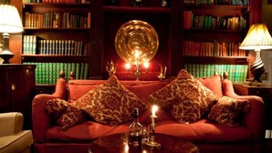 Une pièce chaleureuse avec un canapé rouge, des coussins à motifs, une table ronde sur laquelle se trouvent une bouteille et une bougie, le tout entouré d’étagères remplies de livres.