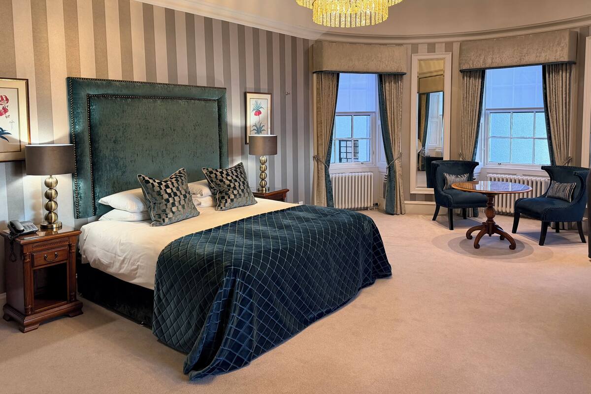 Ein Hotelzimmer mit einem großen Bett, einer Kristallleuchter, einer Sitzecke mit Tisch und Stühlen sowie Blick ins Freie durch große Fenster.