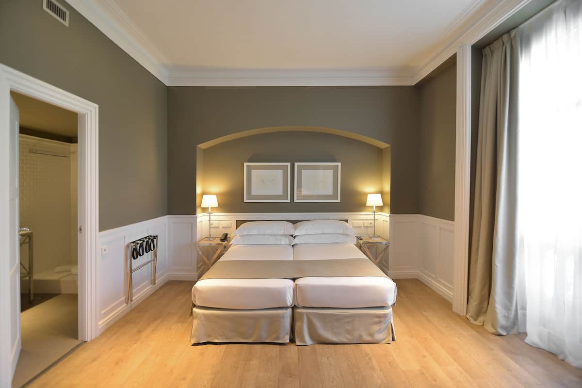 Una cama grande con dos mesitas de noche, un cabecero con dos cuadros enmarcados, y un baño visible a través de una puerta abierta.