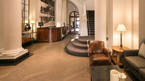 Un vestíbulo amplio con una imponente escalera, una recepción y una sala de estar con una mesa de centro y una lámpara.