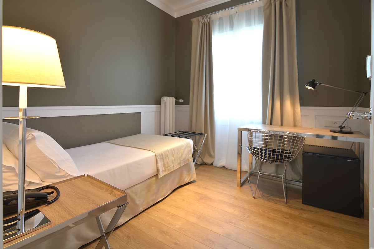 Una habitación de hotel con una cama, un escritorio con silla, una lámpara y un refrigerador pequeño.