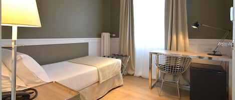 A hotel room with a bed, a desk with a chair, a lamp, and a small refrigerator.