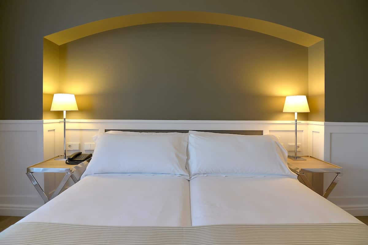 Una cama bien tendida con sábanas blancas, dos lámparas de noche y un teléfono en la mesita.