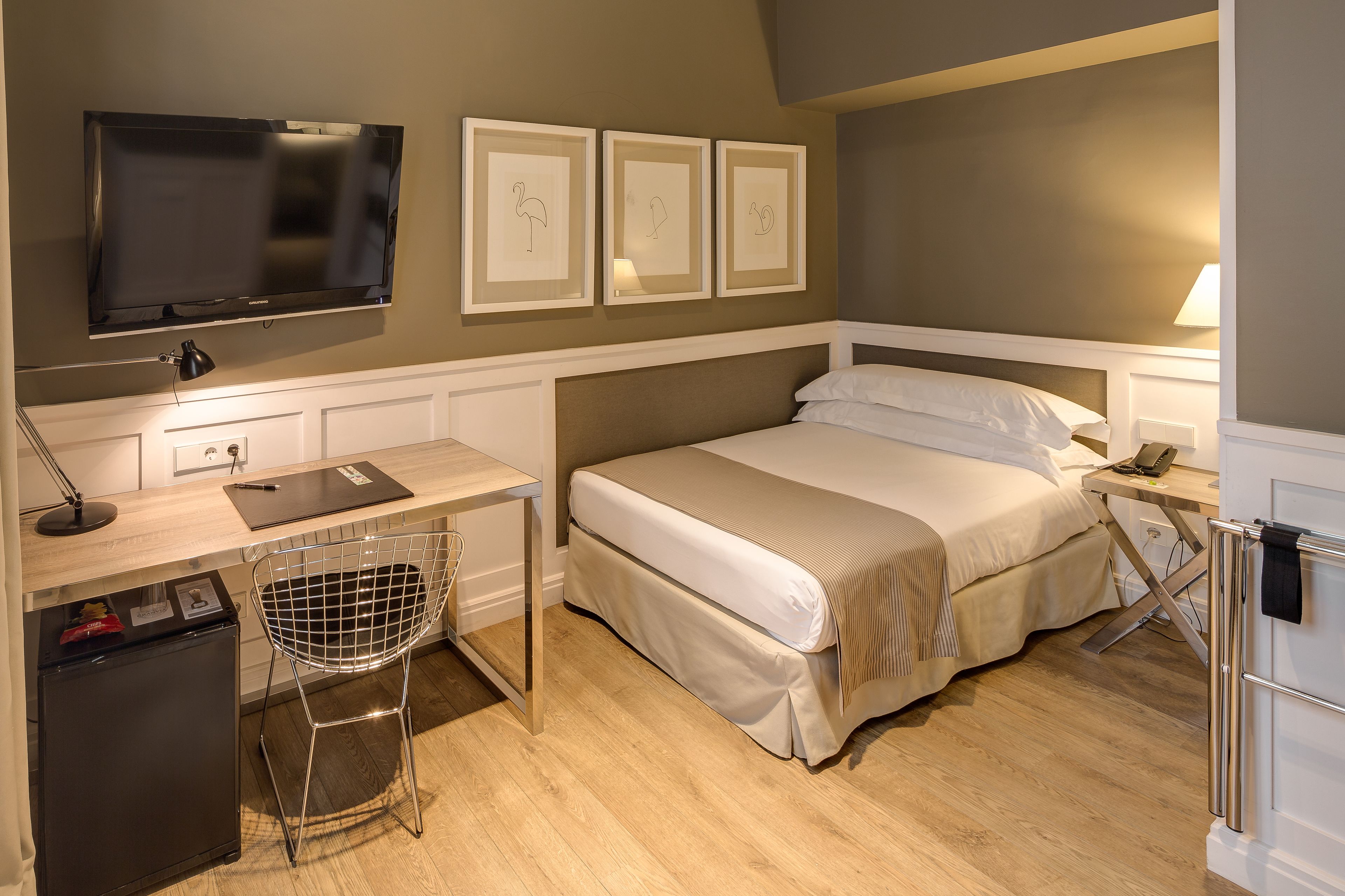 A compact hotel room with a bed, desk, chair, TV, and refrigerator.