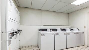 A laundry room with four commercial washers and dryers, a tiled floor, and a white ceiling with fluorescent lighting.