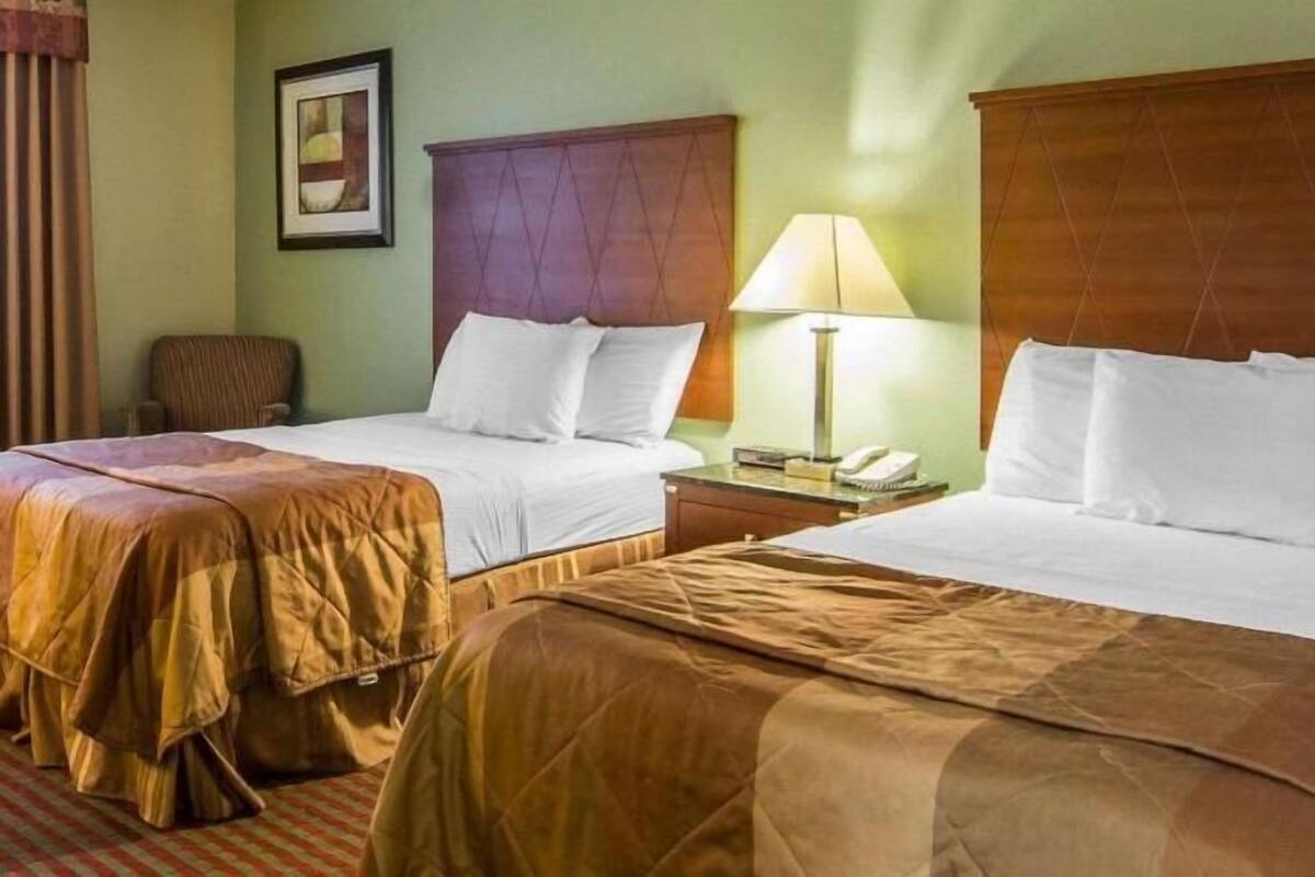 Habitación de hotel con dos camas, una mesita de noche con lámpara, una silla y un cuadro en la pared.