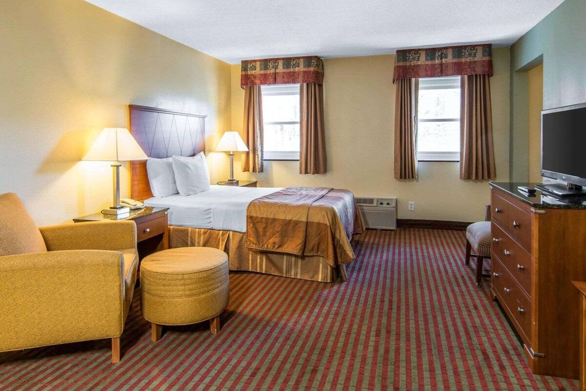 Una habitación de hotel con una cama, dos lámparas de noche, un televisor, una silla, un aparador y piso alfombrado.