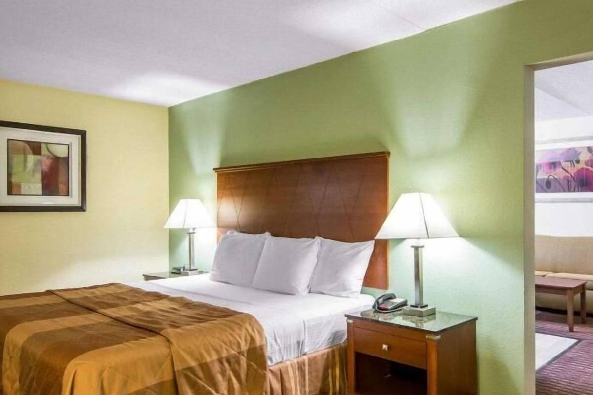 Una habitación de hotel con una cama, dos mesitas de noche con lámparas, un cabecero de madera y un sofá en la habitación contigua.