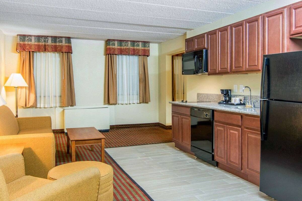 Una habitación de hotel compacta con kitchenette, un sofá, una pequeña mesa y un ventanal con cortinas.