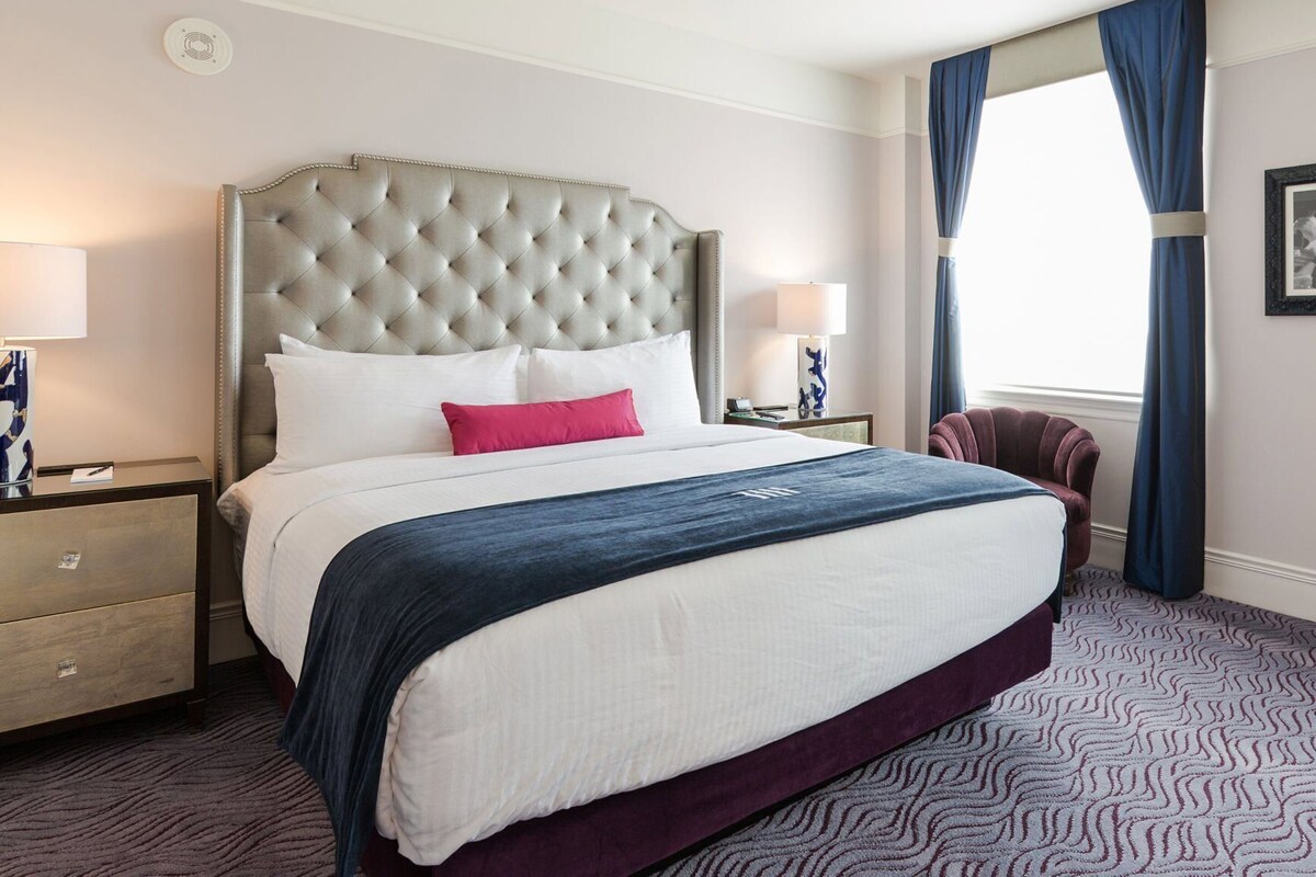 A hotel room with a tufted headboard, a large bed with white and dark blue bedding, a nightstand with a lamp, and a window with blue curtains.