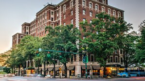 A large brick building with multiple stories, likely a hotel, featuring a prominent entrance with a canopy and a street view with traffic lights and trees.