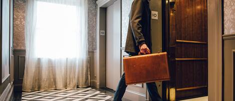 A man carrying a briefcase walking towards an elevator in a hotel lobby with patterned flooring and a large window with sheer curtains.