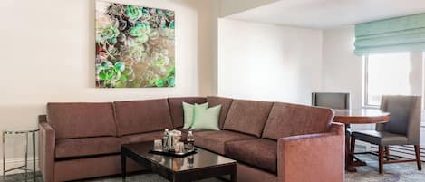 A living room with a brown sectional sofa, a coffee table, a painting on the wall, a dining table with chairs, and a large window with a view.