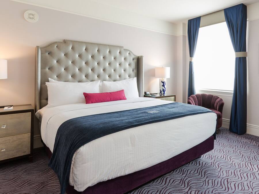 A hotel room with a large bed, a headboard, a nightstand, a chair, and a window with curtains.
