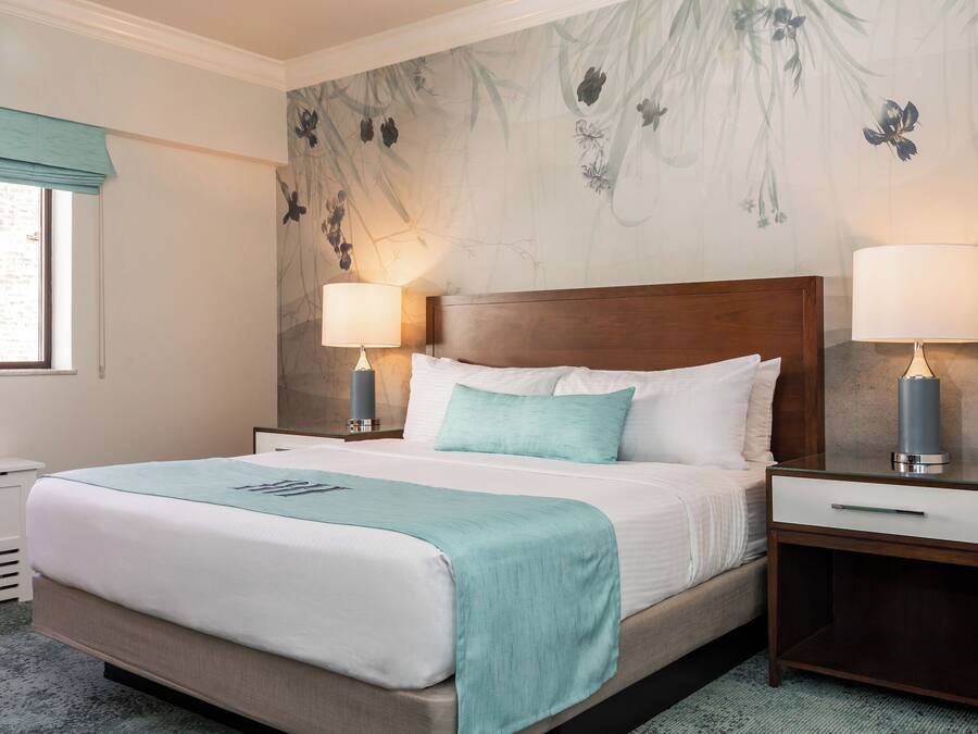 Een opgemaakt bed met een blauwgroene plaid, twee bedlampjes en een nachtkastje met een lade.