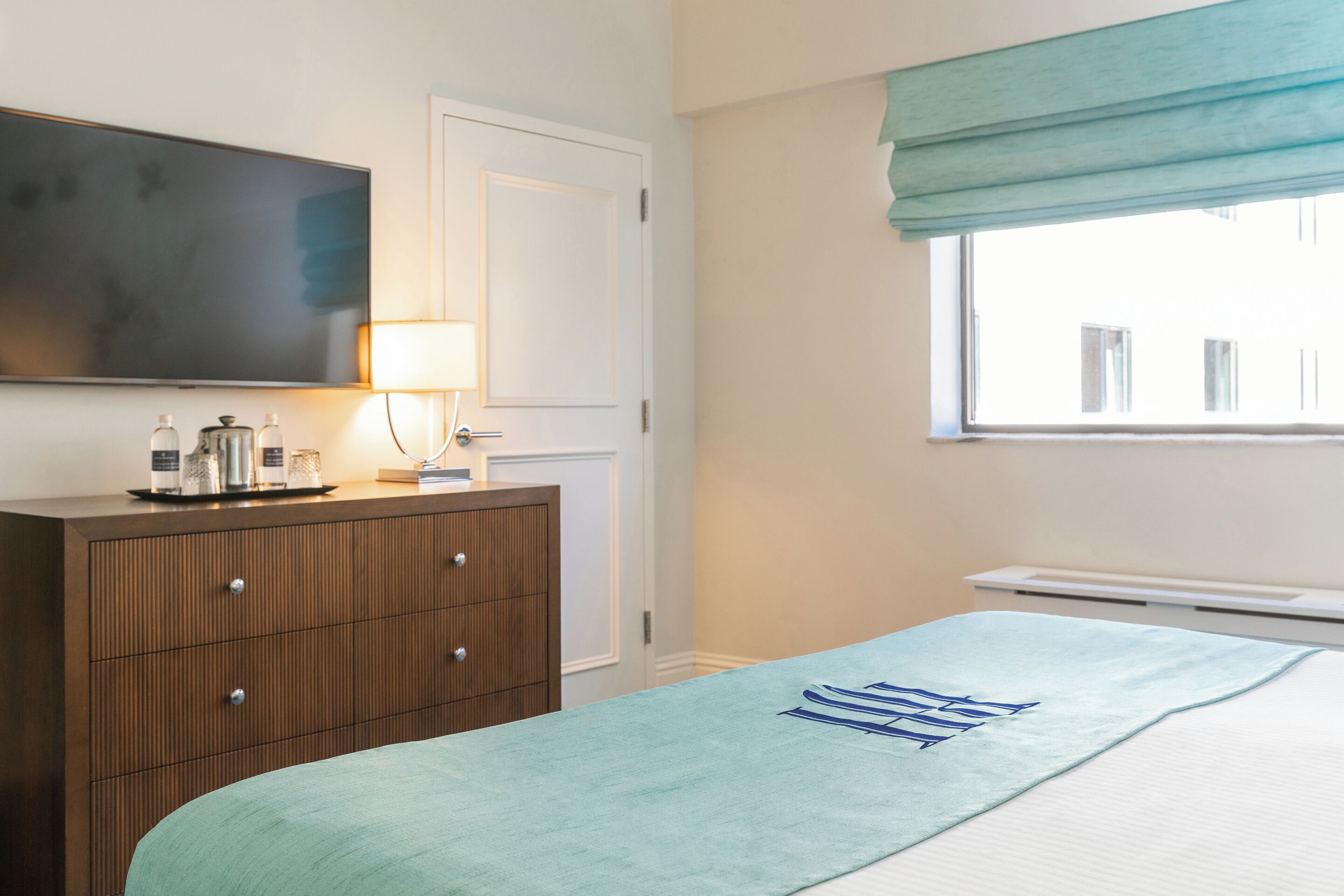 Une chambre à coucher comprenant un lit, une commode, un téléviseur et une fenêtre donnant sur des immeubles.