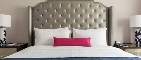 Habitación de hotel con cabecera capitonada, ropa de cama blanca, una manta azul y una almohada roja.
