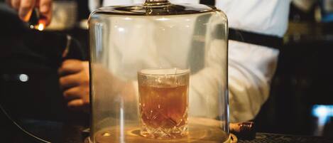 A glass of amber liquid under a glass dome on a wooden coaster.
