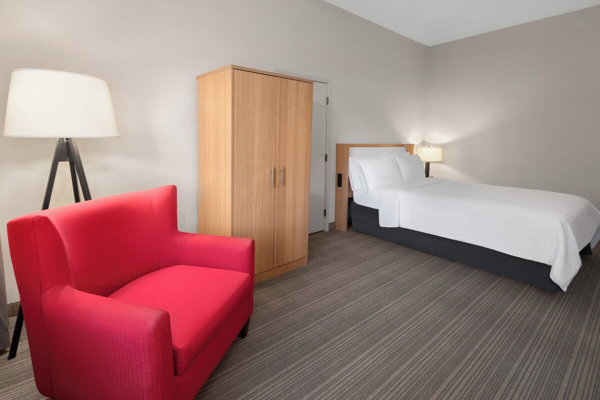 Quarto de hotel com poltrona vermelha, guarda-roupa de madeira, cama com roupa de cama branca e abajur.