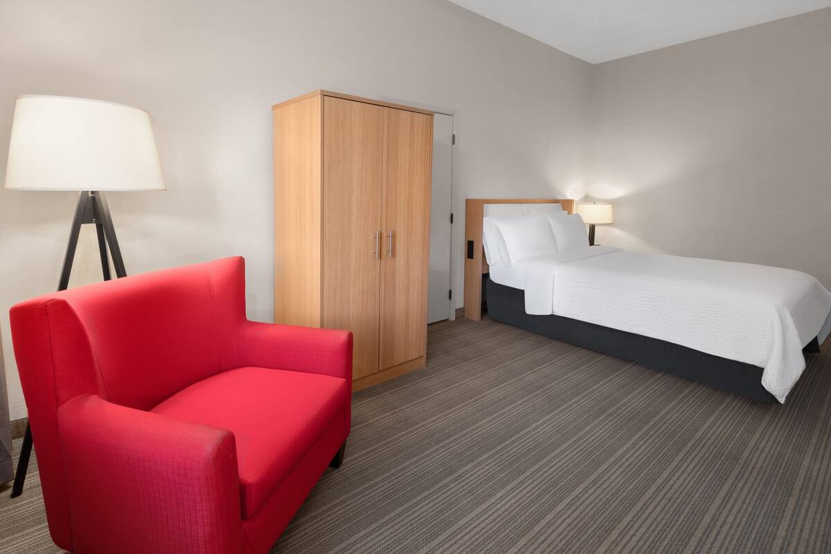 Ein Hotelzimmer mit einem roten Sessel, einem Holzschrank, einem Bett mit weißer Bettwäsche und einer Stehlampe.