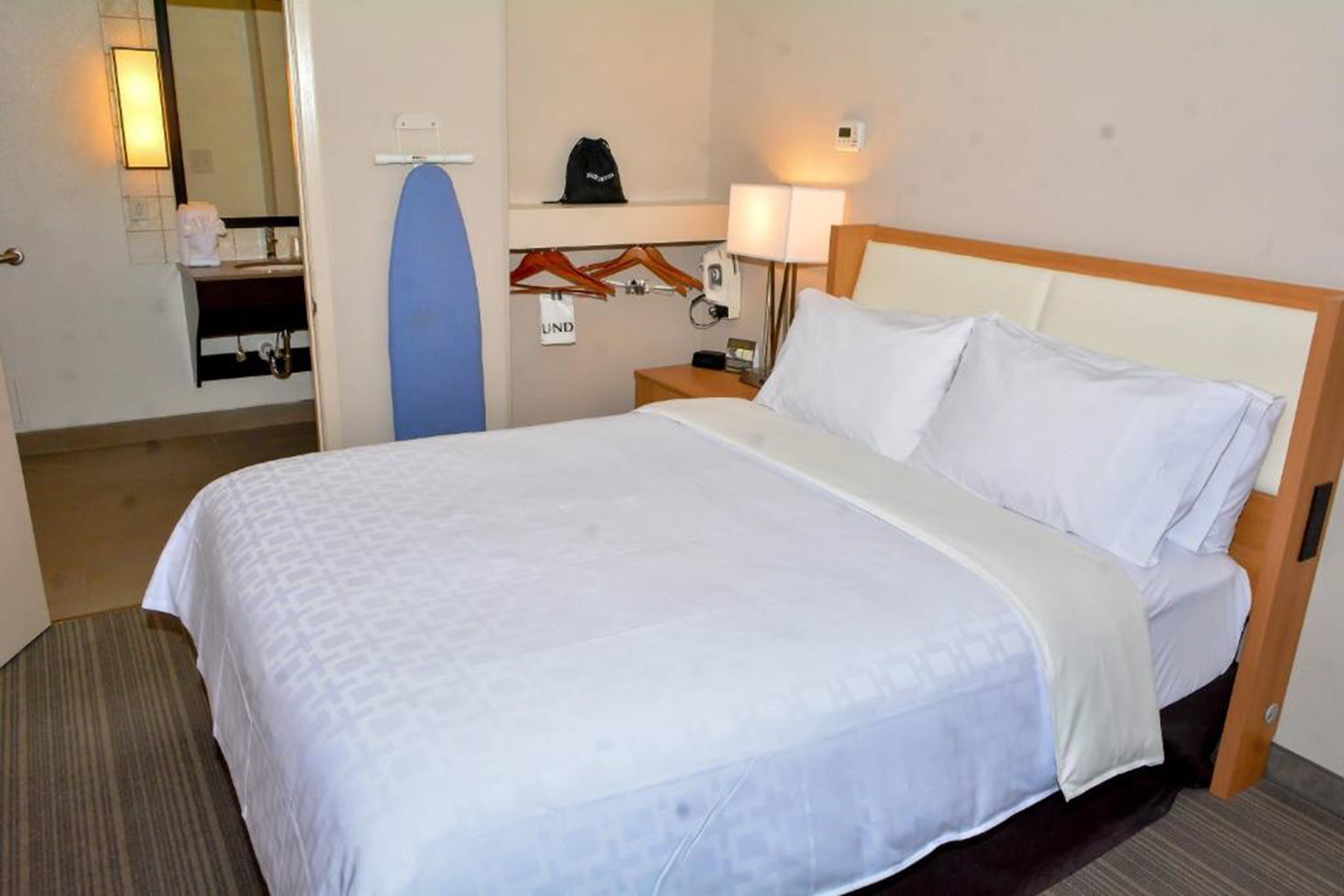 A hotel room with a bed, a bedside table with a lamp, a mirror, and a coat rack.