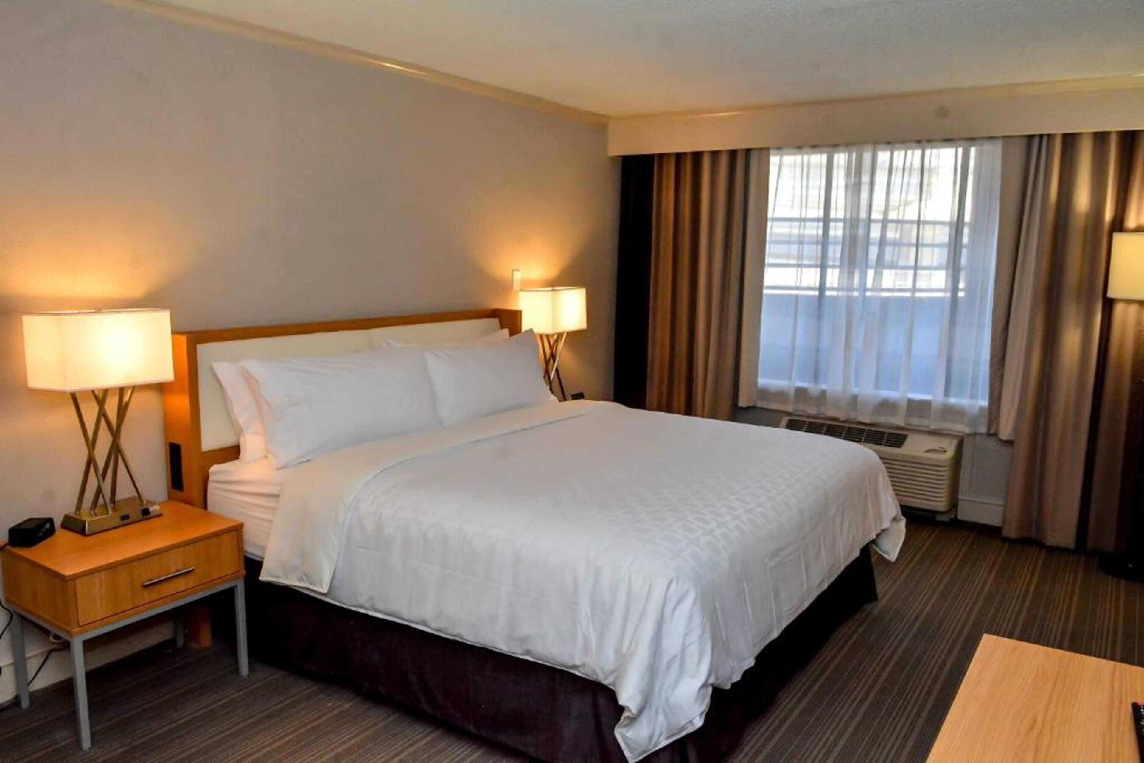 Ein Hotelzimmer mit einem großen Bett, zwei Nachttischlampen, einem Nachttisch, einem Fenster mit Vorhängen und einer Klimaanlage.