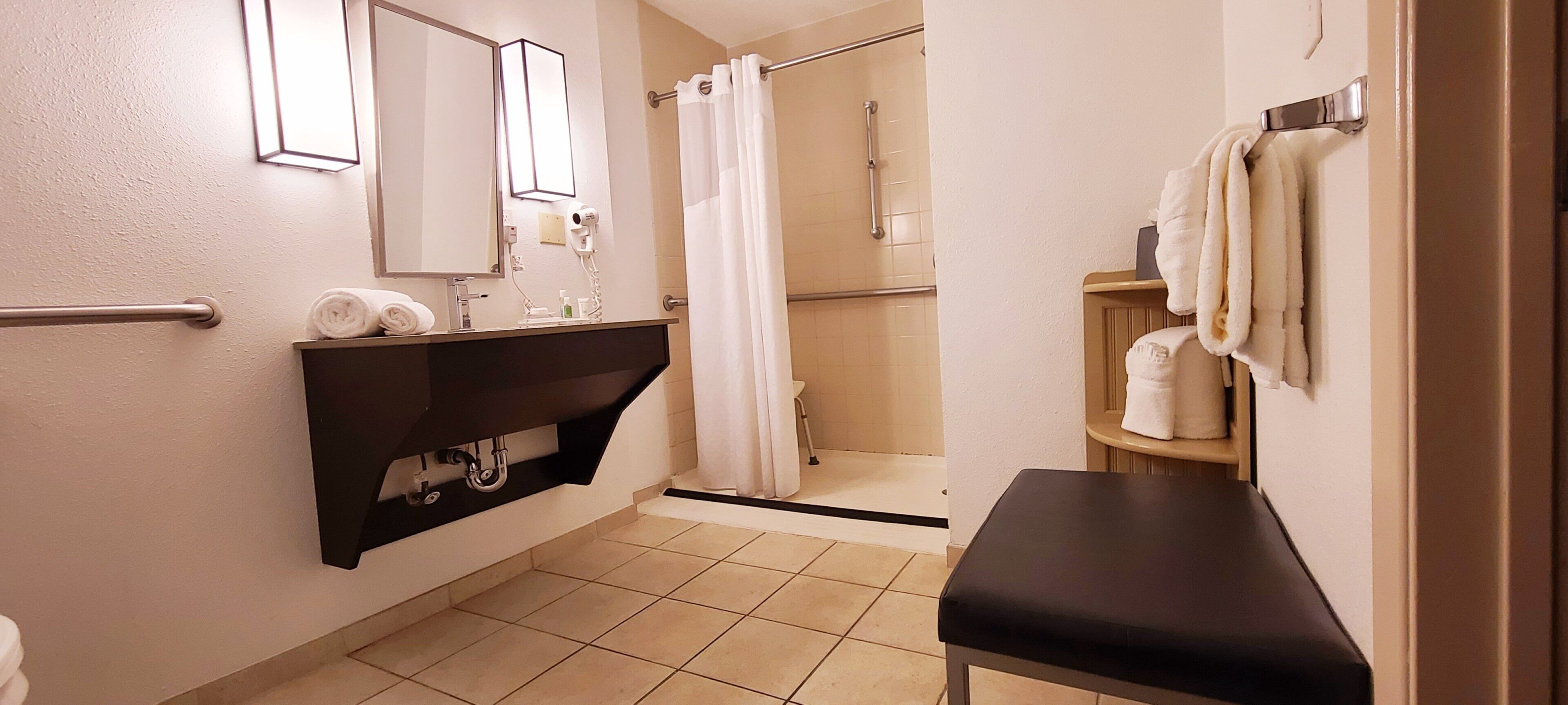 A bathroom with a sink, mirror, towel rack, shower area, and a bench.