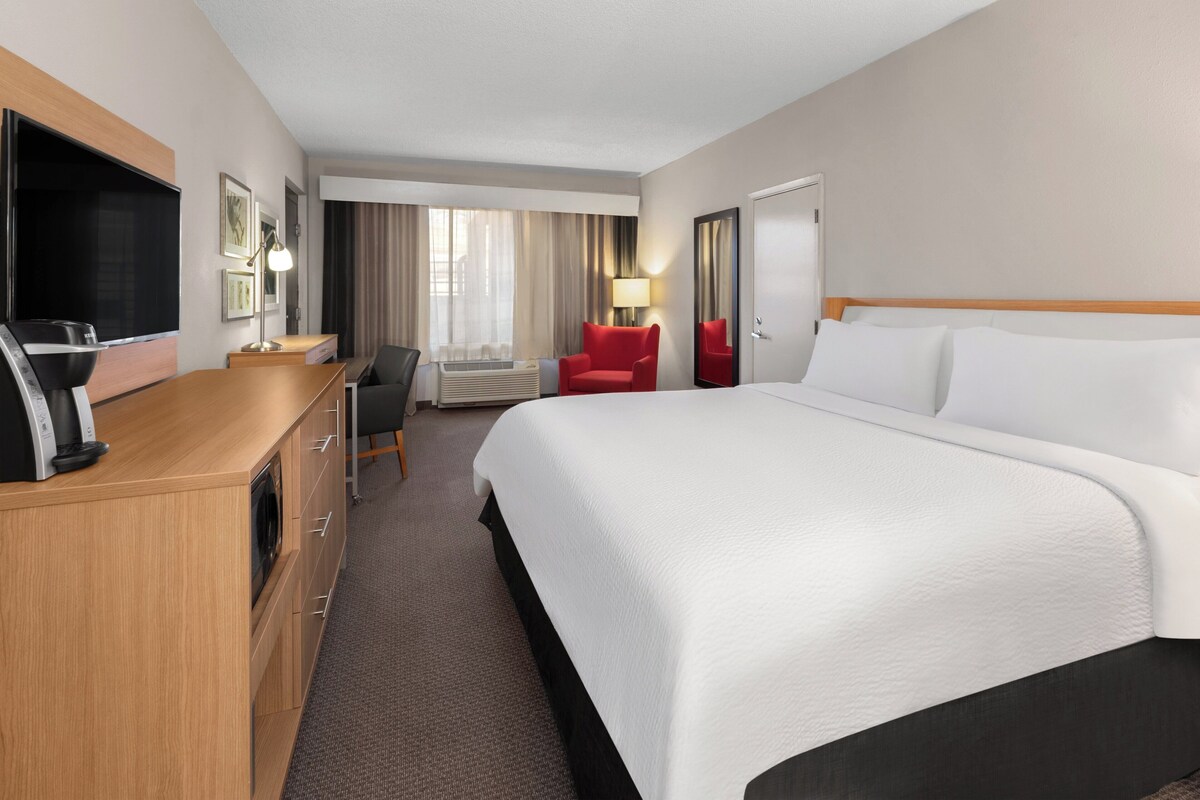 Quarto de hotel com uma cama grande, uma escrivaninha com televisão de tela plana, uma poltrona vermelha e um abajur.