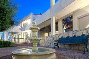 Un patio con una fuente de piedra, una banca y un edificio con amplios ventanales.