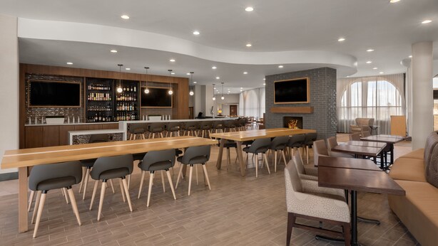 Een moderne bar/loungeruimte met een lange houten bar, diverse zitplaatsen zoals barstoelen en fauteuils, een open haard en een aan de muur bevestigde televisie.