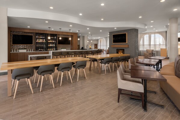 Un bar/salon moderne doté d’un long comptoir en bois, de nombreux espaces pour s’asseoir, notamment des tabourets et des chaises de bar, d’une cheminée et d’un téléviseur fixé au mur.